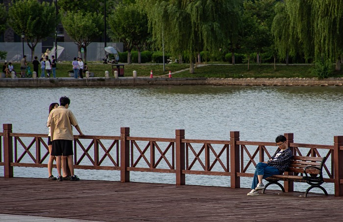 A man and woman stand on a wooden pier, while another man sits on a bench, all overlooking a body of water.  (Captioned by AI)